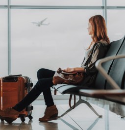 Tranquil woman is sitting on bench with luggage in lounge zone. She is looking through window at flying plane while waiting for her flight