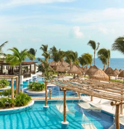 A luxurious tropical resort with a serene blue pool, surrounded by lush palm trees, thatched-roof cabanas, and hammocks. In the background, there's a calming view of the ocean under a clear blue sky.