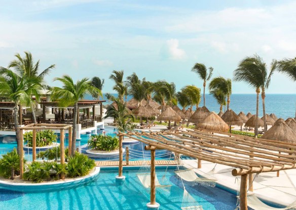 A luxurious tropical resort with a serene blue pool, surrounded by lush palm trees, thatched-roof cabanas, and hammocks. In the background, there's a calming view of the ocean under a clear blue sky.